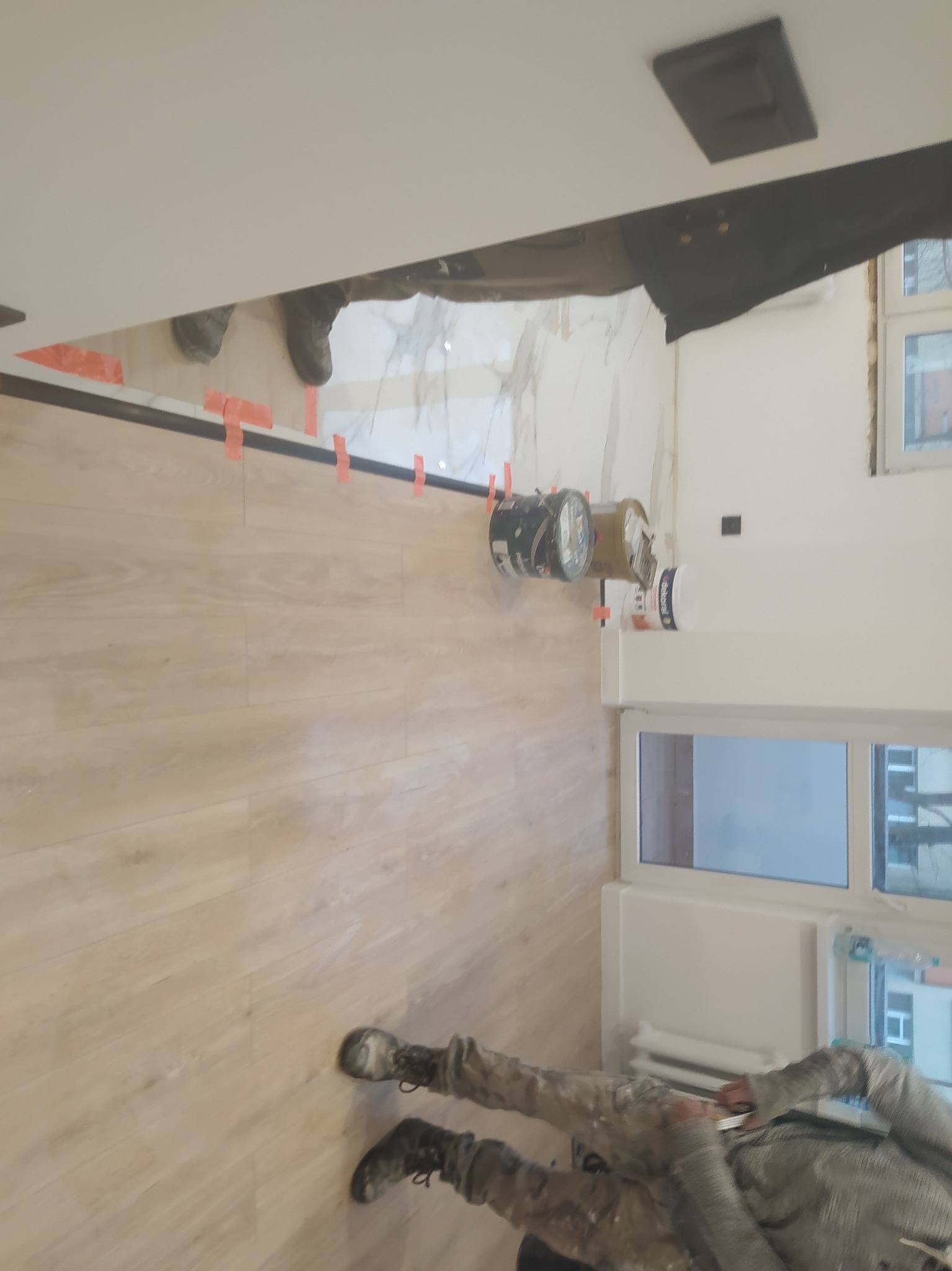 Podczas remontu: świeżo ułożone jasne panele podłogowe, zabezpieczone taśmą, kontrastują z marmurowymi płytkami na ścianie. Widoczne puszki z farbą i pracownik w ubraniu roboczym.