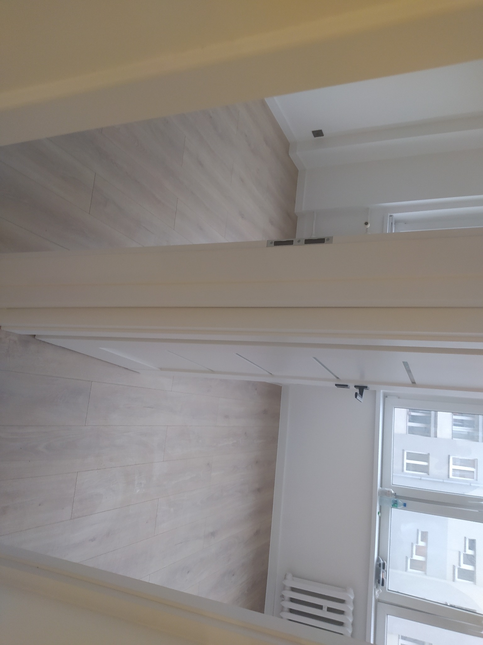 Widok z góry na jasne panele podłogowe ułożone wzdłuż ściany z oknem, widoczny fragment białej ramy drzwiowej i grzejnika.