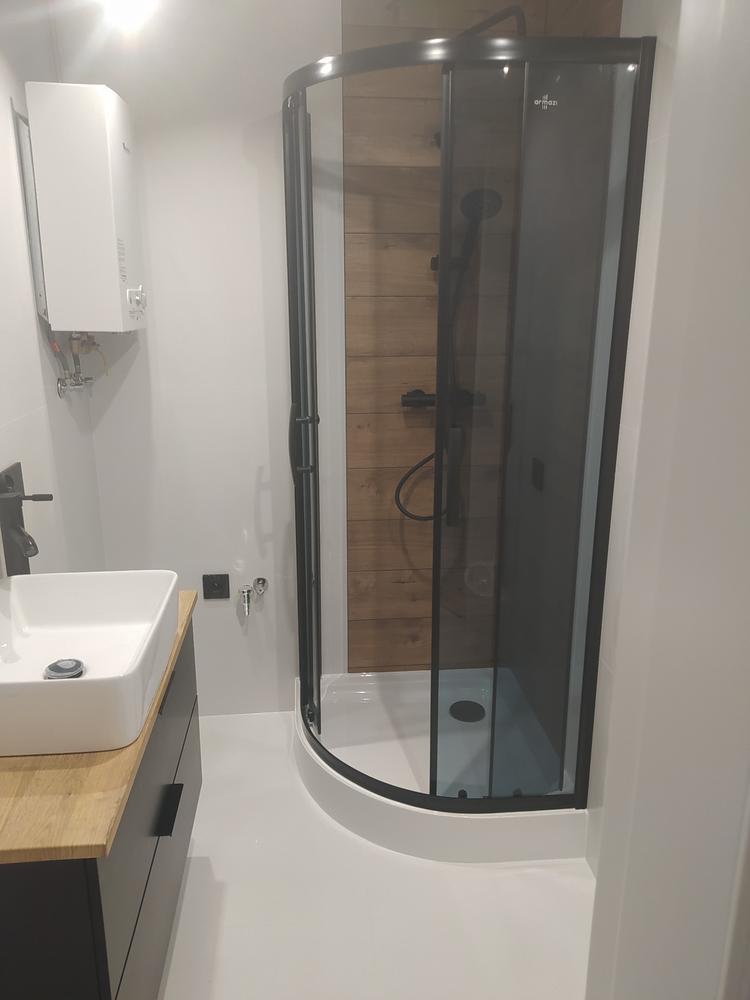 Nowoczesna łazienka z kabiną prysznicową z czarnymi profilami i drewnianym panelem dekoracyjnym wewnątrz, białą umywalką nablatową z czarną armaturą i grzejnikiem.