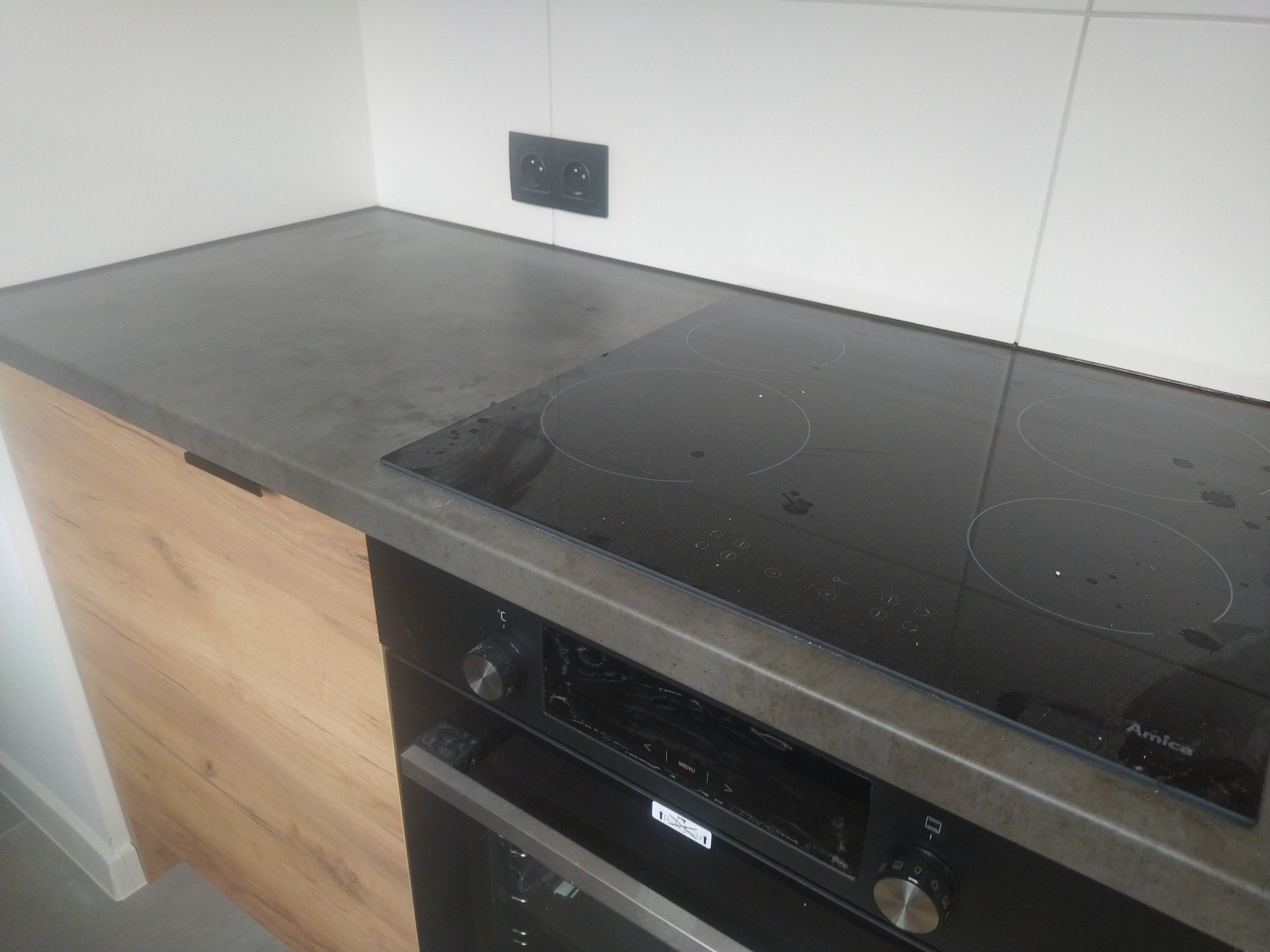 Fragment kuchni z drewnianą szafką, szarym blatem, czarną płytą indukcyjną Amica i piekarnikiem, widoczne białe płytki na ścianie i podwójne gniazdko elektryczne.
