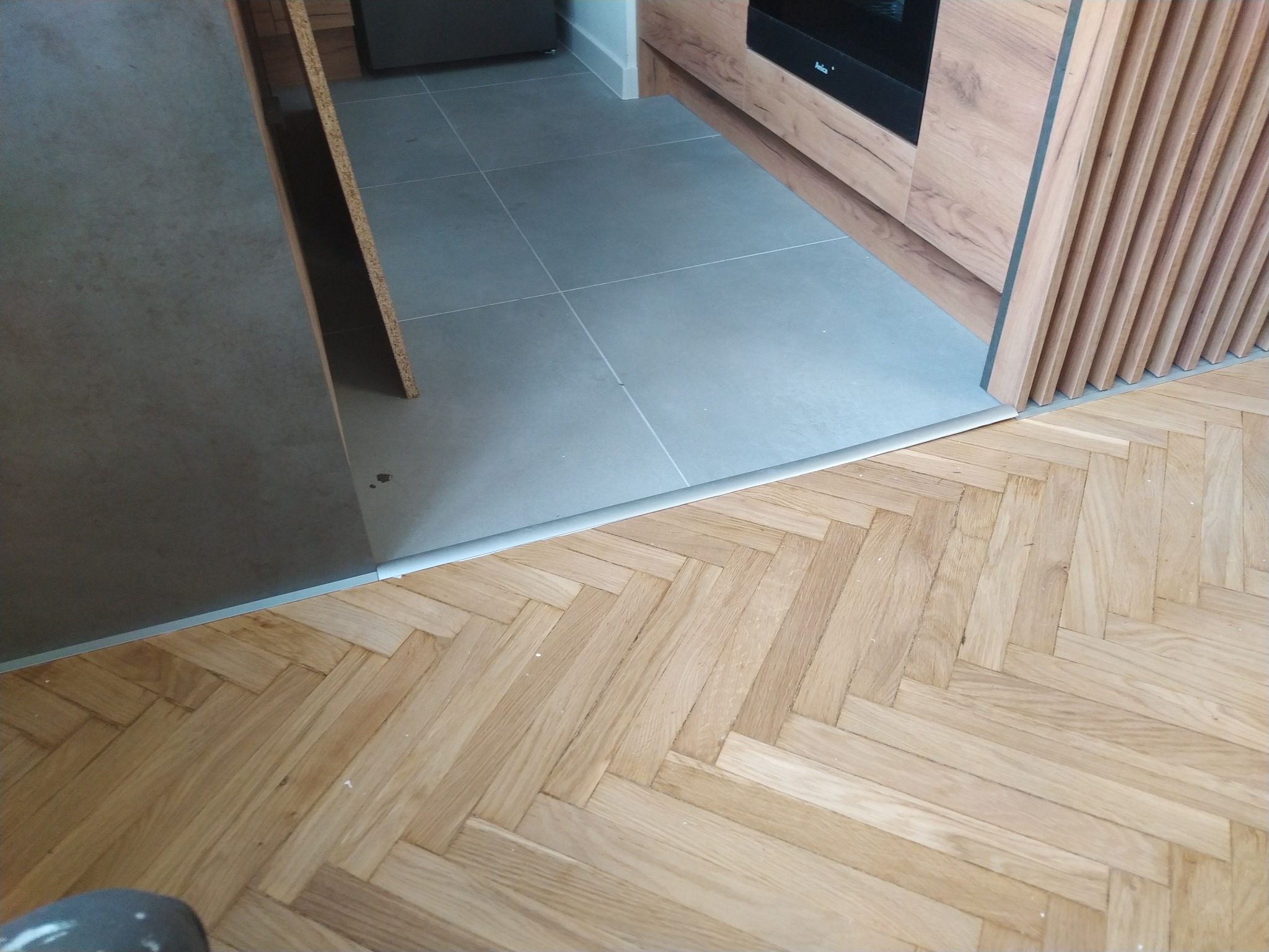 Połączenie jasnej, parkietowej podłogi ułożonej w jodełkę z szarą płytką podłogową w kuchni, widoczna listwa progowa i fragment zabudowy kuchennej z drewnianymi lamelami.