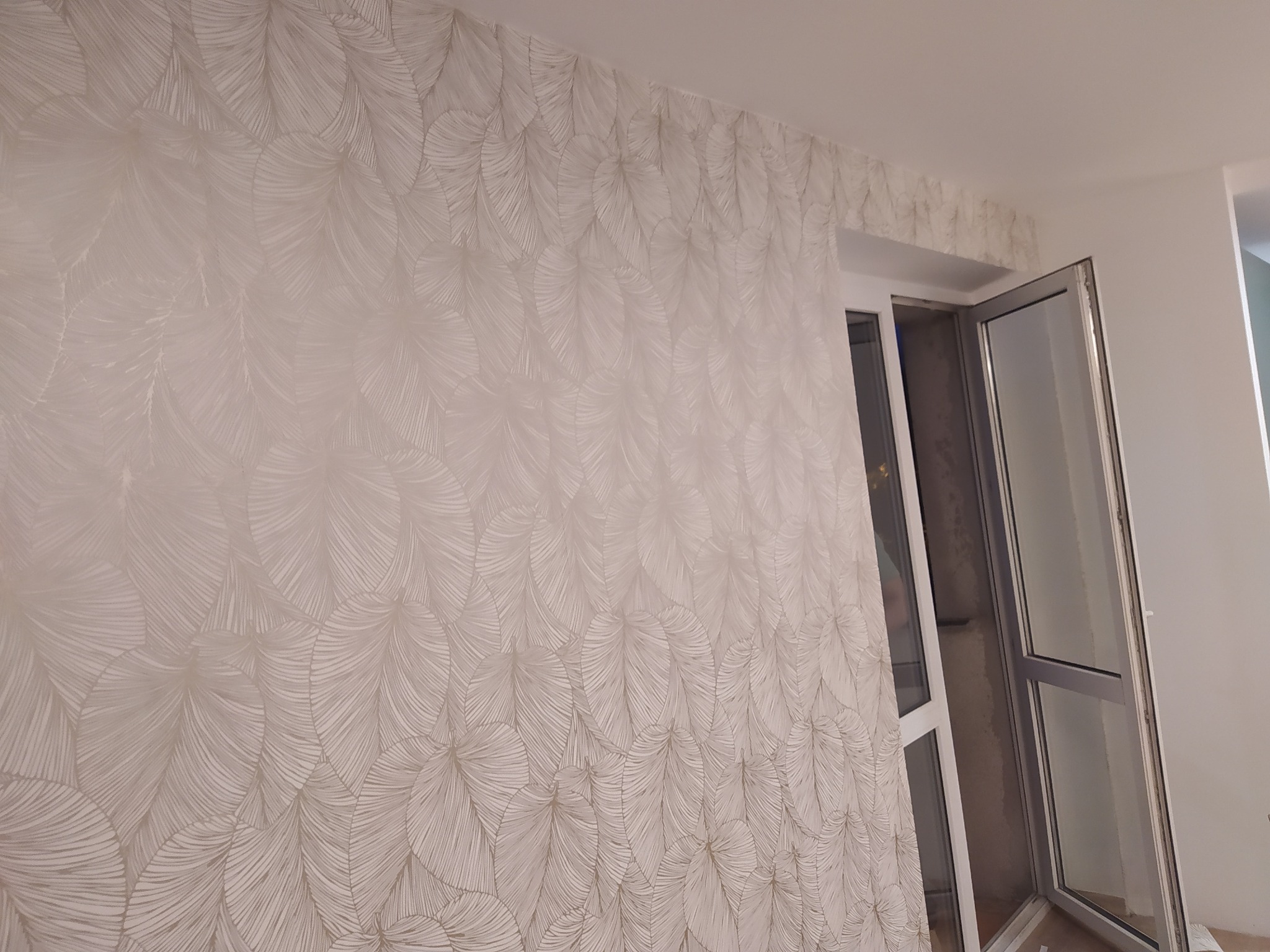 Ściana pokryta tapetą w delikatny, piórkowy wzór, z fragmentem niezakończonego montażu na górze, przylegająca do otwartego okna balkonowego z białymi ramami.