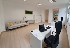 Jasne, przestronne biuro z białymi ścianami i drewnianą podłogą. W tle sofa z żółtą poduszką, biurko z lampką i komoda. Na pierwszym planie biurko z laptopem, notatkami i czarnym krzesłem biurowym.