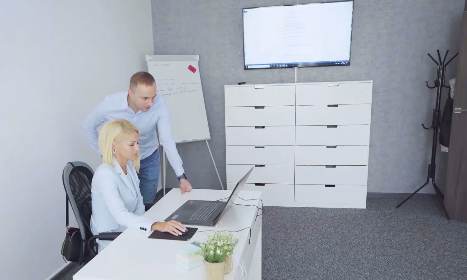 Kobieta pracuje na laptopie przy biurku, mężczyzna stoi obok i patrzy na ekran, w tle flipchart z zapiskami i monitor na szafce.