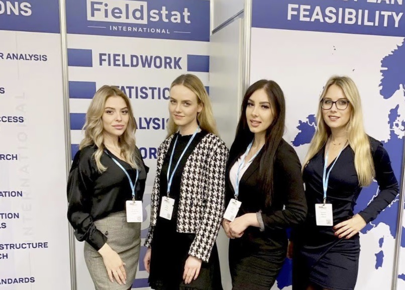 Cztery kobiety w strojach biznesowych prezentują się na tle stoiska targowego Fieldstat International z hasłami Fieldwork, Statistic Analysis i Feasibility.