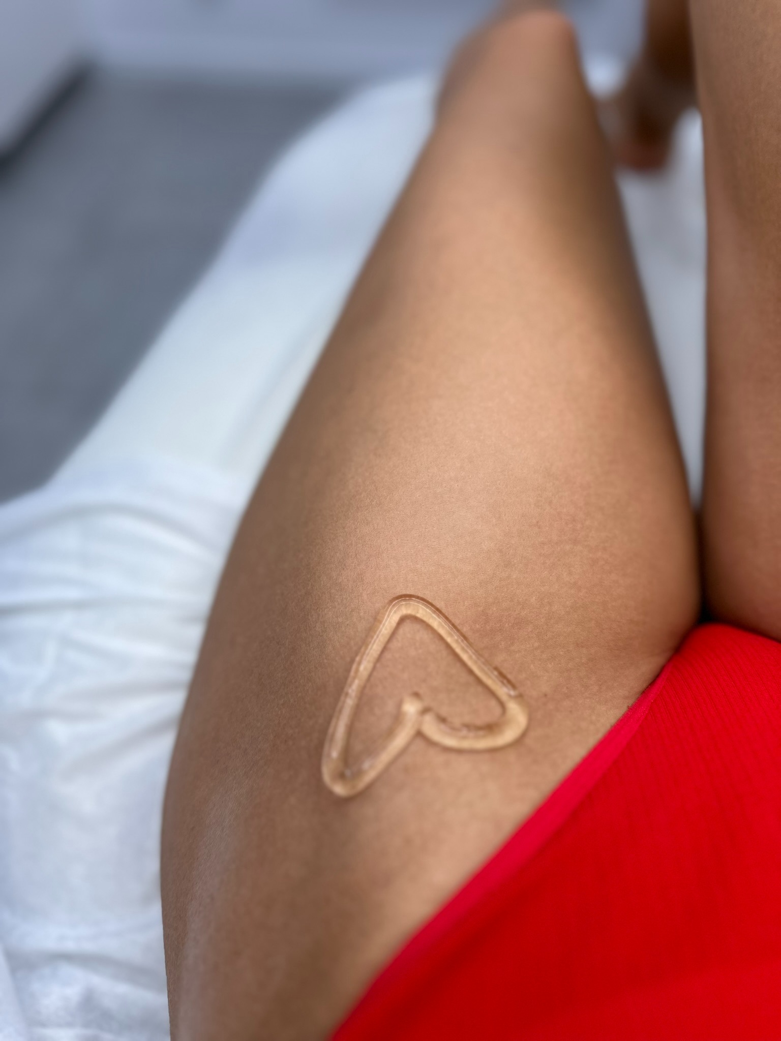 Krem w kształcie serca na skórze uda, widoczny fragment czerwonej bielizny, białe prześcieradło w tle.