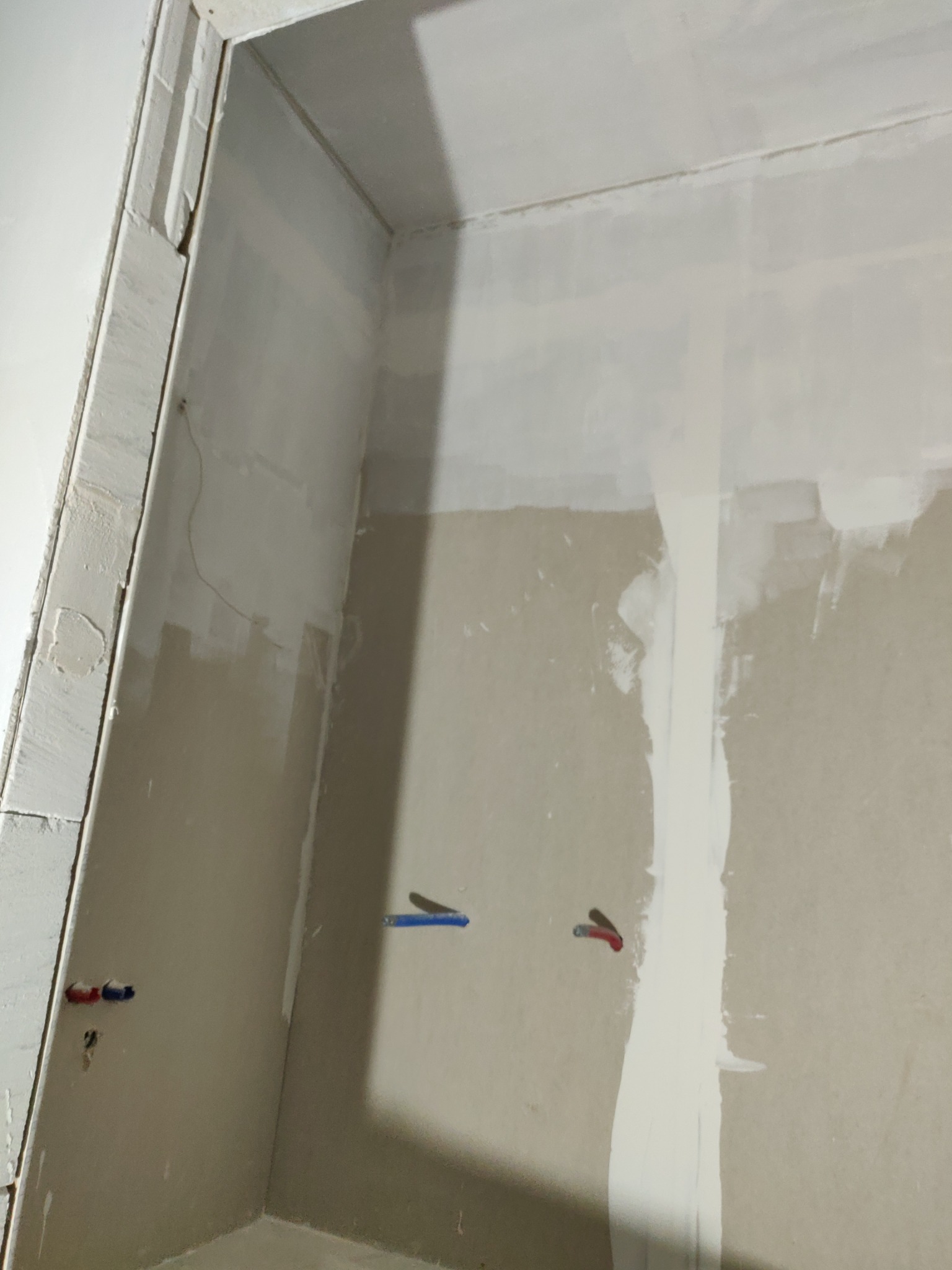 Surowa ściana w trakcie szpachlowania, widoczne ślady po szpachli, niebieskie i czerwone rurki instalacyjne wystające ze ściany