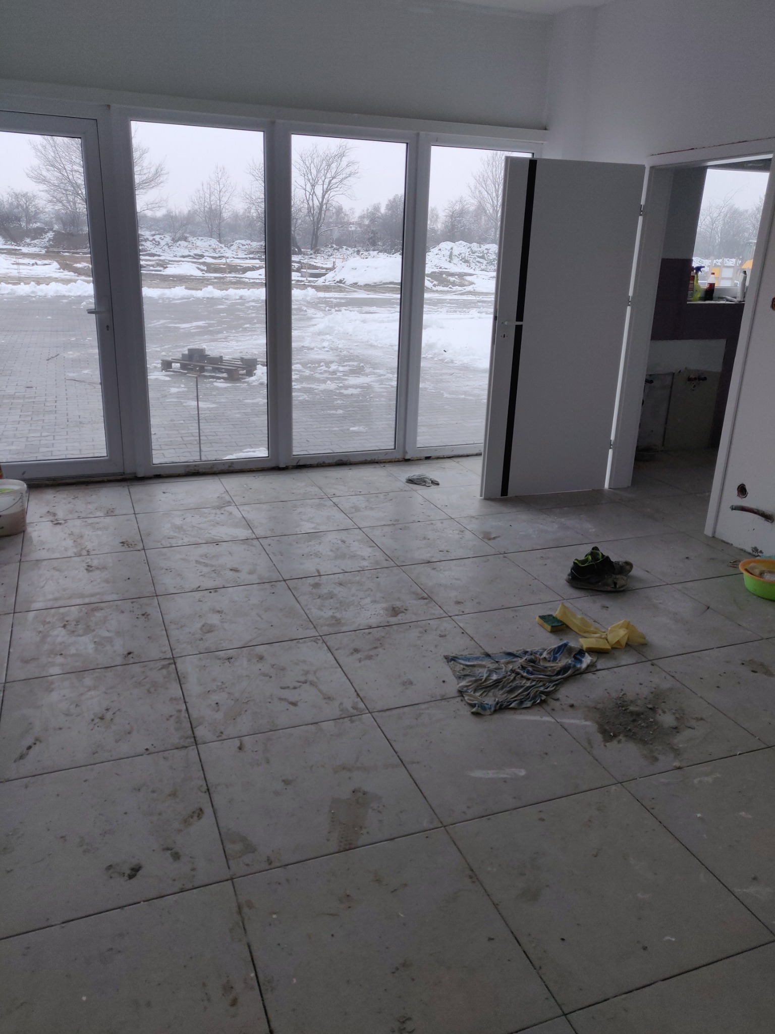 Podłoga w trakcie czyszczenia po remoncie, widoczne ślady zabrudzeń, szmata, gąbki i buty robocze, w tle duże okna z widokiem na zimowy krajobraz.