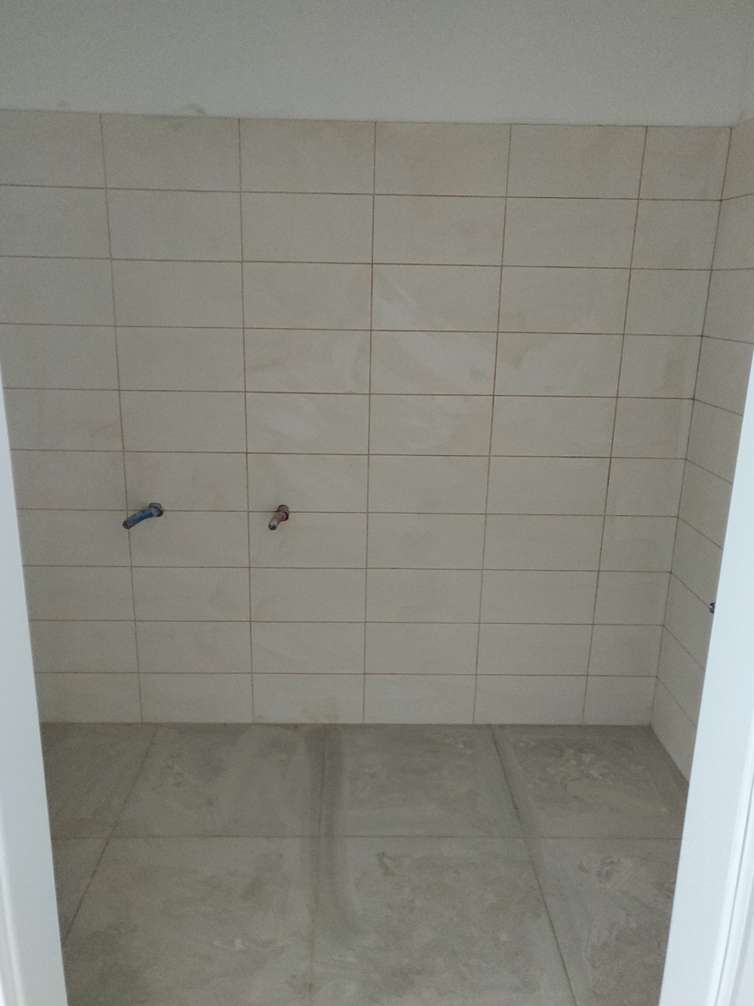 Wnętrze łazienki w trakcie prac wykończeniowych: beżowe płytki na ścianach i podłodze, widoczne wyprowadzenia instalacji wodnej, ujęcie zza futryny drzwi.