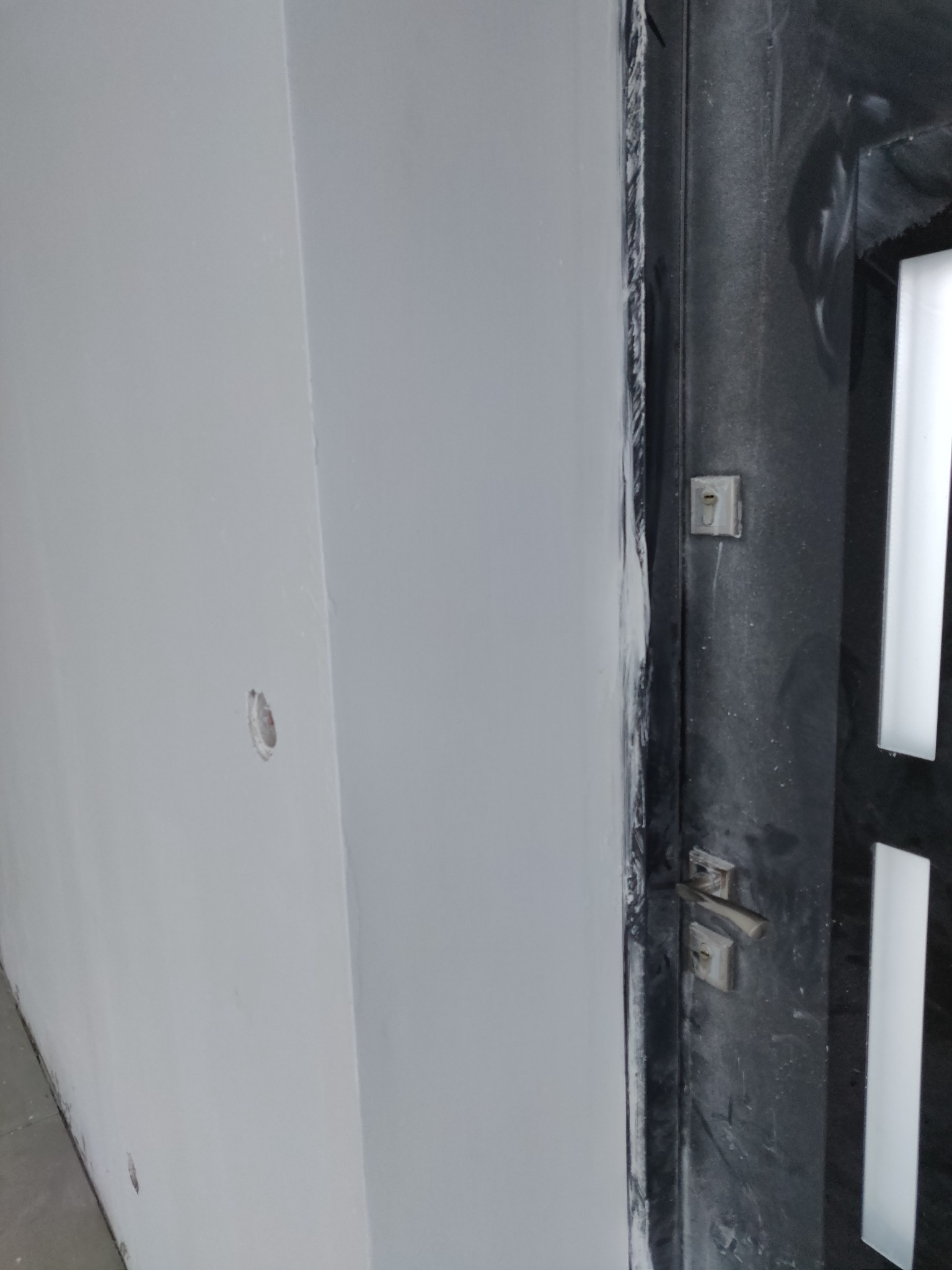 Świeżo pomalowana na szaro ściana z widocznym otworem na gniazdko elektryczne, sąsiadująca z czarną, częściowo zaklejoną taśmą malarską ościeżnicą drzwi z metalową klamką i zamkiem