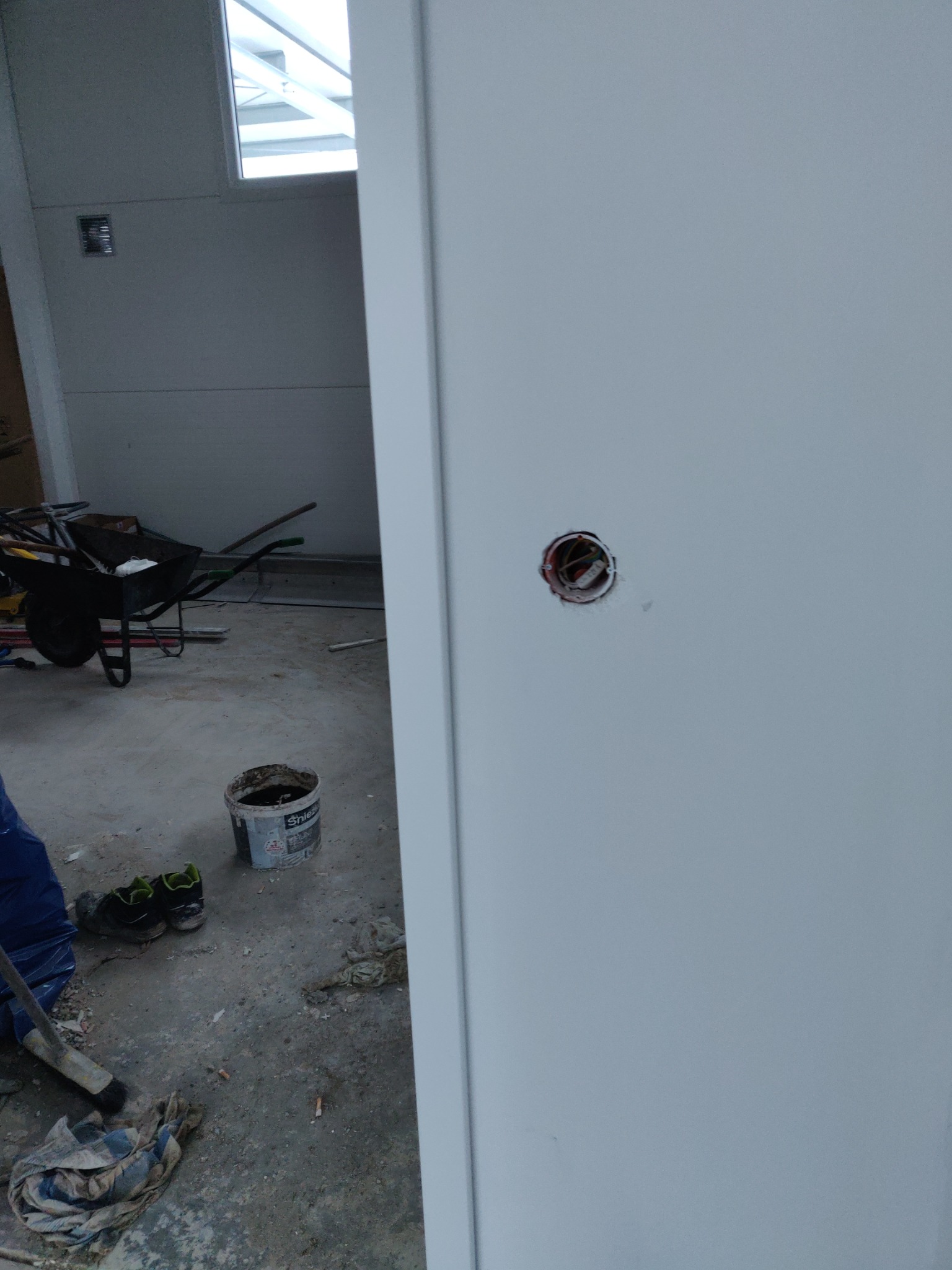 Biała ściana z otworem na puszkę elektryczną w trakcie prac wykończeniowych, widoczny fragment wózka budowlanego, wiadro po farbie i robocze buty.