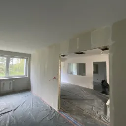 Wnętrze w trakcie remontu: ściany przygotowane do malowania, zabezpieczona podłoga folią, widoczne profile narożnikowe i fragment okna z zielenią na zewnątrz.