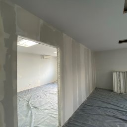 REMPROF - Wnętrze w trakcie prac wykończeniowych: pionowe pasy świeżo nałożonej masy na ścianach z płyt kartonowo-gipsowych, podłoga zabezpieczona folią malarską, widoczna rama drzwiowa i fragment sufitu...