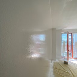 REMPROF - Świeżo pomalowane na biało wnętrze z widoczną fakturą na ścianie, zabezpieczona folią podłoga, otwarte okno balkonowe wpuszczające jasne światło.