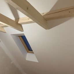 REMPROF - Poddasze po remoncie: drewniane belki stropowe, okno dachowe w wykończonej wnęce i świeżo pomalowane ściany w odcieniach bieli.