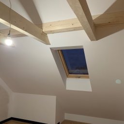 REMPROF - Poddasze z oknem dachowym po remoncie, świeżo pomalowane na biało, widoczne drewniane belki stropowe i fragment muru z cegły po prawej stronie. Jedna żarówka zawieszona na kablu oświetla przestrzeń.