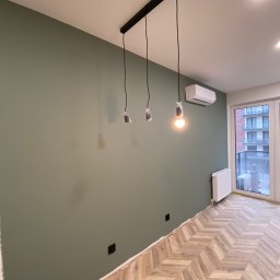 REMPROF - Pomieszczenie po malowaniu ścian na oliwkowy kolor, widoczne niedokończone oświetlenie z trzema żarówkami, grzejnik, klimatyzator i parkiet ułożony w jodełkę.