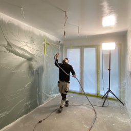 REMPROF - Pracownik w masce ochronnej aplikuje farbę natryskową w pomieszczeniu zabezpieczonym folią malarską, widoczna lampa na statywie oświetlająca ścianę.