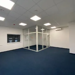 Puste biuro po remoncie z niebieską wykładziną, przeszkloną ścianką działową i białymi ścianami, oświetlone kwadratowymi panelami LED w suficie.