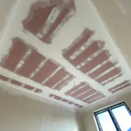 Sufit z widocznymi płytami kartonowo-gipsowymi, częściowo pokrytymi białą masą szpachlową, w trakcie prac wykończeniowych. W tle widoczne okno z niebieskim niebem.