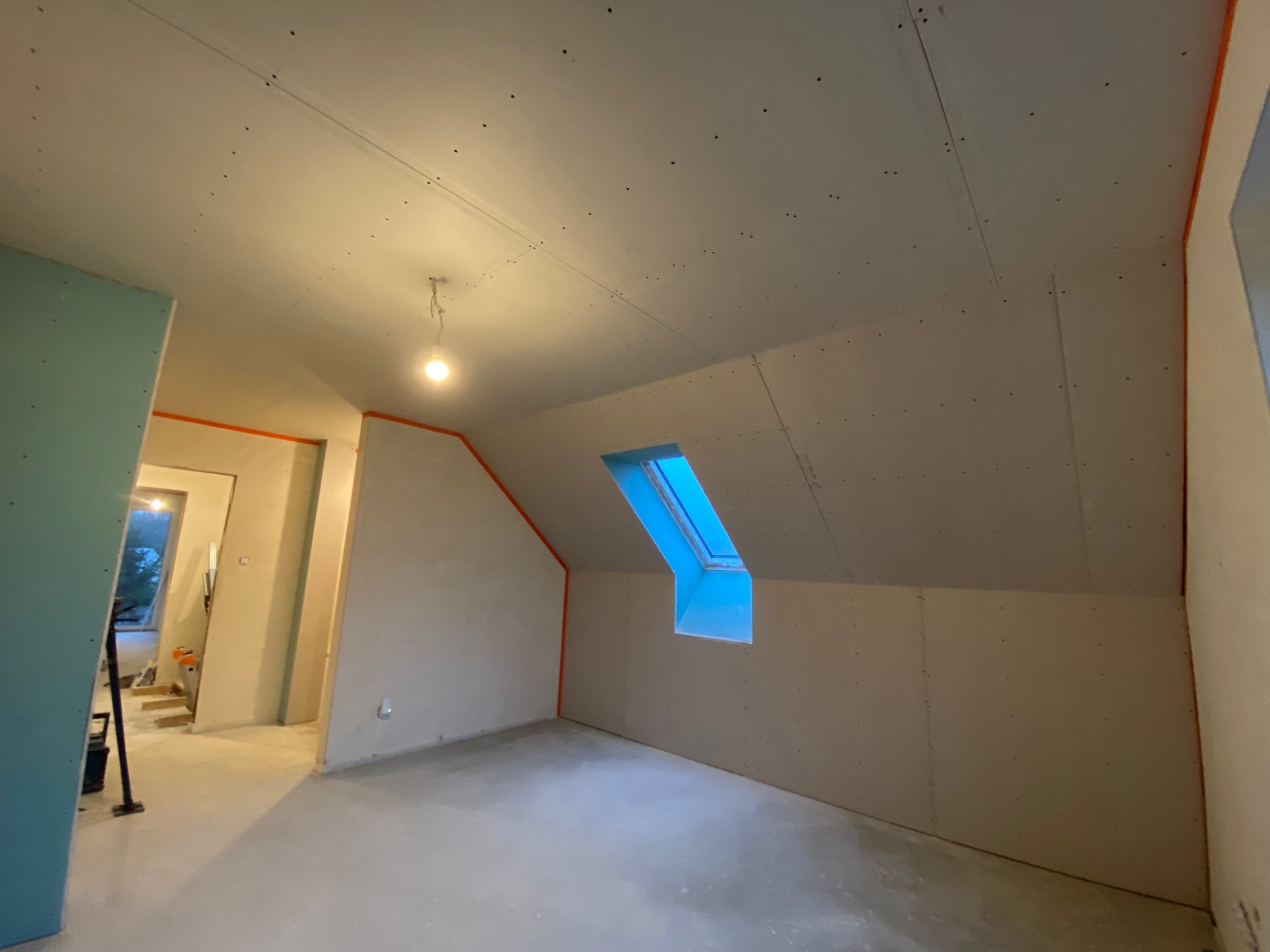 Poddasze w trakcie prac wykończeniowych: widoczna konstrukcja ze skosami, okno dachowe wpuszczające jasne światło, płyty kartonowo-gipsowe zamontowane na ścianach i suficie, ślady po wkrętach...