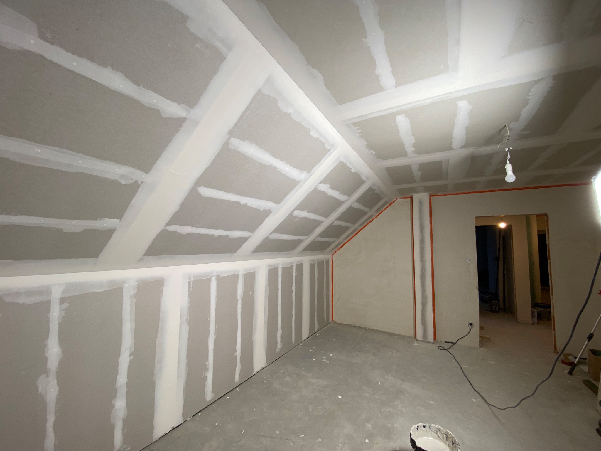 Poddasze w trakcie wykańczania: widoczne płyty kartonowo-gipsowe na skosach dachu i ścianach, łączenia płyt zaszpachlowane, surowy beton na podłodze, wisząca żarówka.