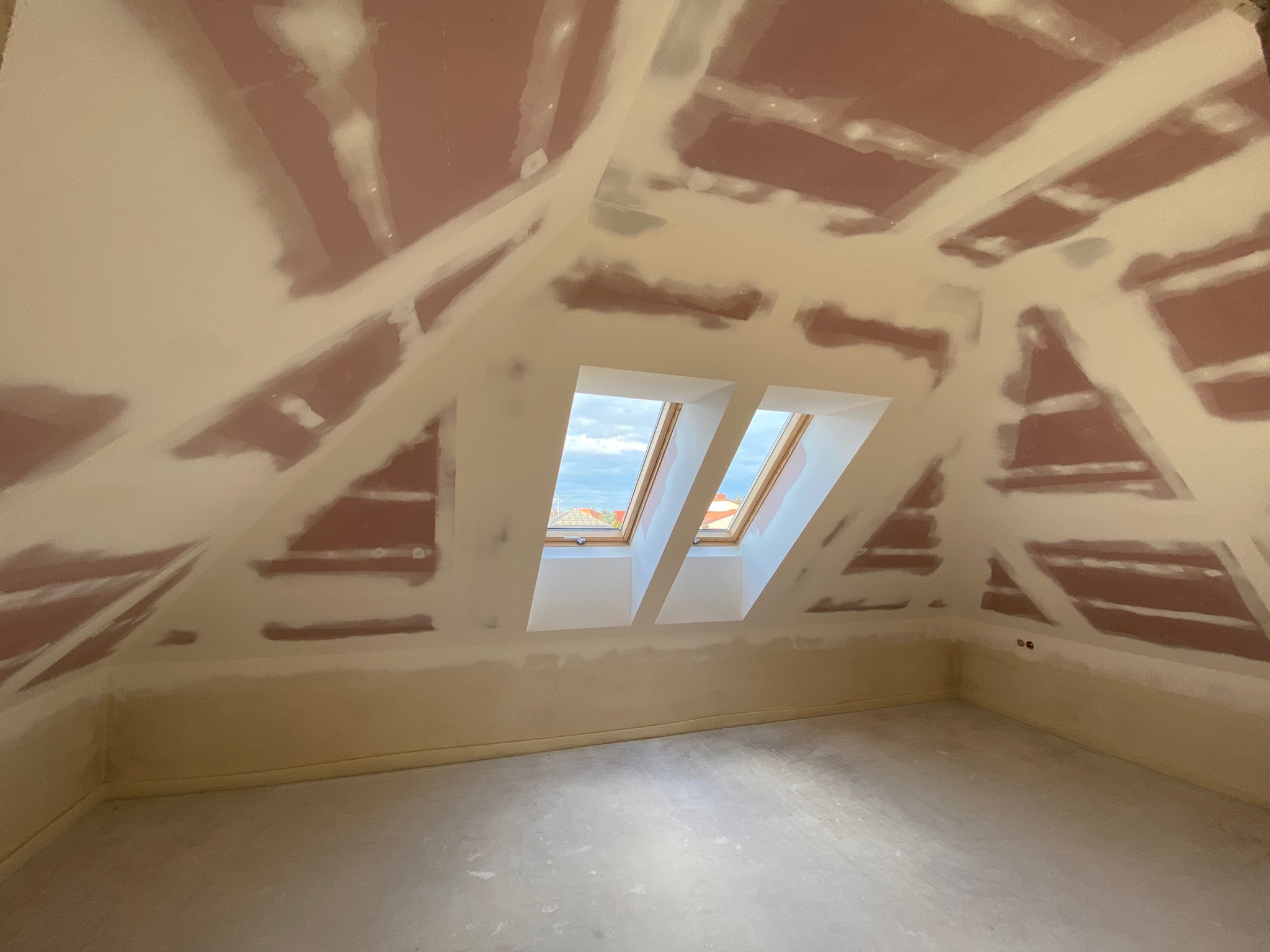 Poddasze ze skośnym sufitem w trakcie prac wykończeniowych, widoczne fragmenty niezamaskowanej płyty kartonowo-gipsowej i nałożona warstwa szpachli, w tle okno dachowe z widokiem na niebo.