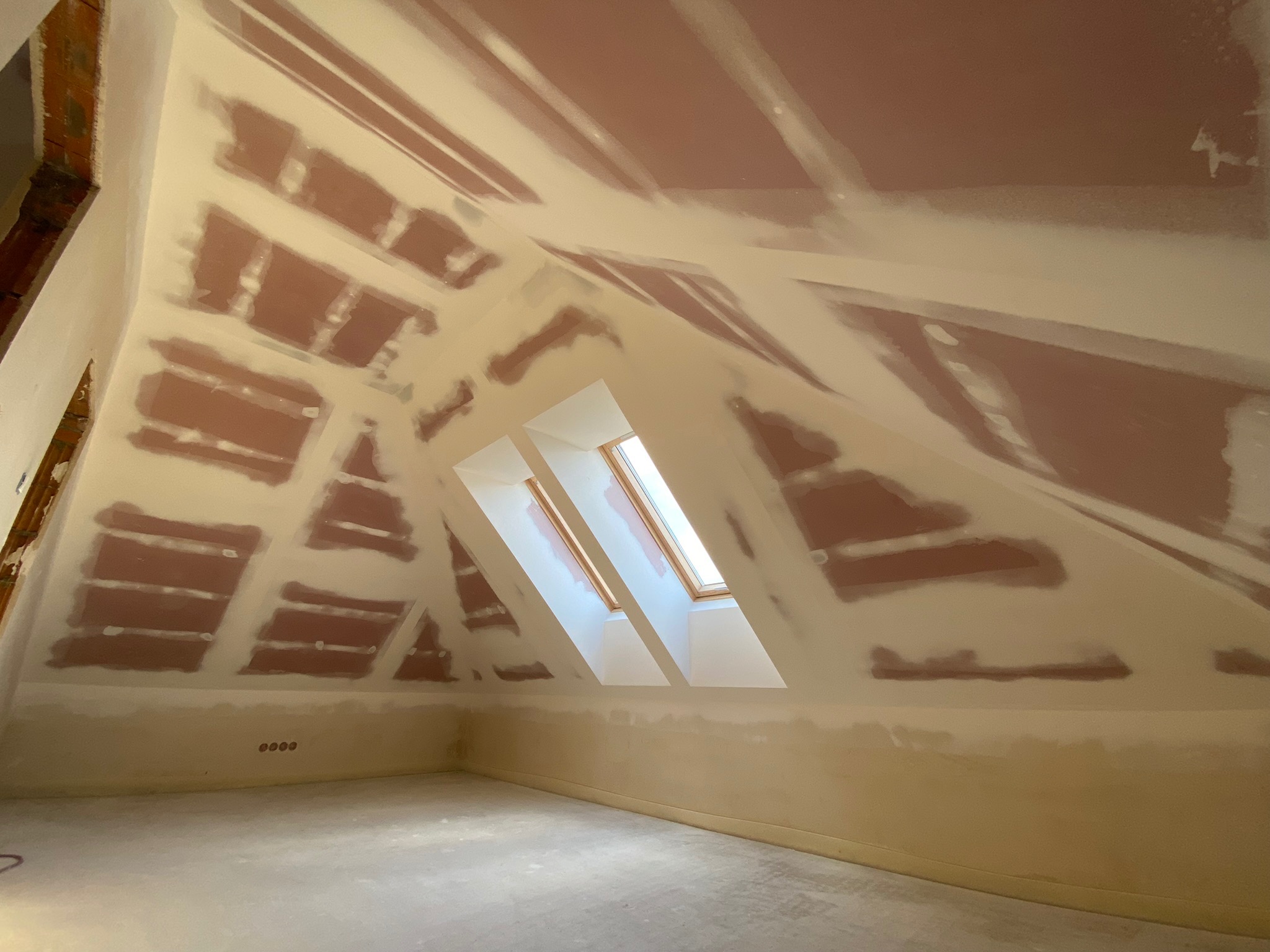 Poddasze z oknami dachowymi w trakcie prac wykończeniowych; widoczne fragmenty płyt gipsowo-kartonowych pokryte warstwą szpachli, przygotowane do dalszej obróbki.