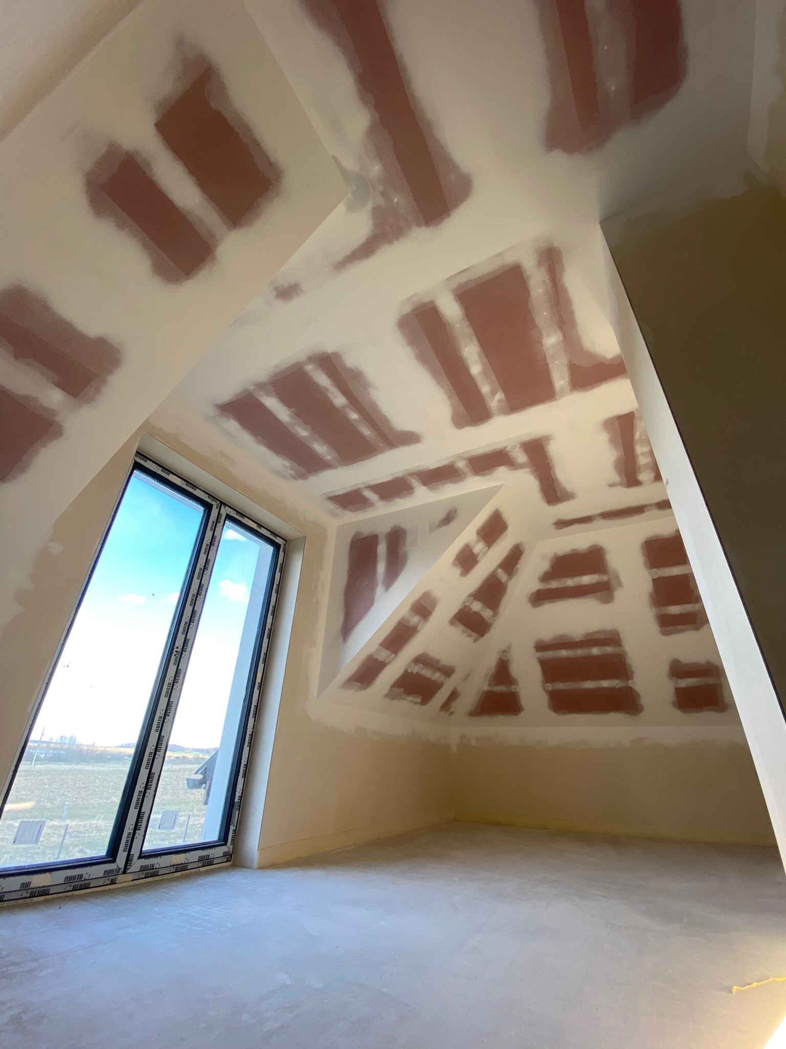 Poddasze w trakcie przygotowania do malowania: widoczne fragmenty zagruntowanej powierzchni na suficie skośnym i ścianach, surowe wykończenie podłogi, duże okno z widokiem na krajobraz.