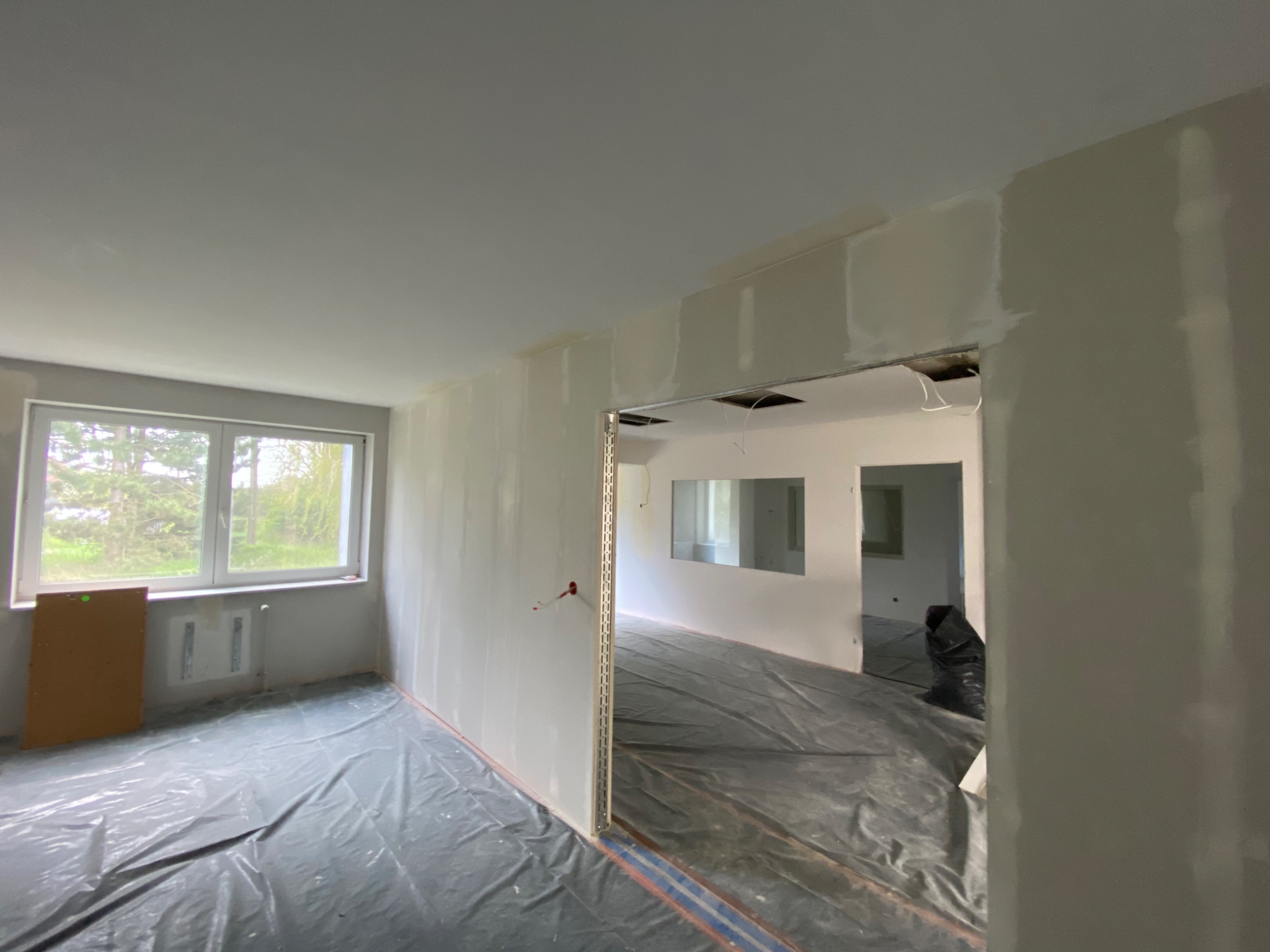 Wnętrze w trakcie remontu: ściany przygotowane do malowania, zabezpieczona podłoga folią, widoczne profile narożnikowe i fragment okna z zielenią na zewnątrz.