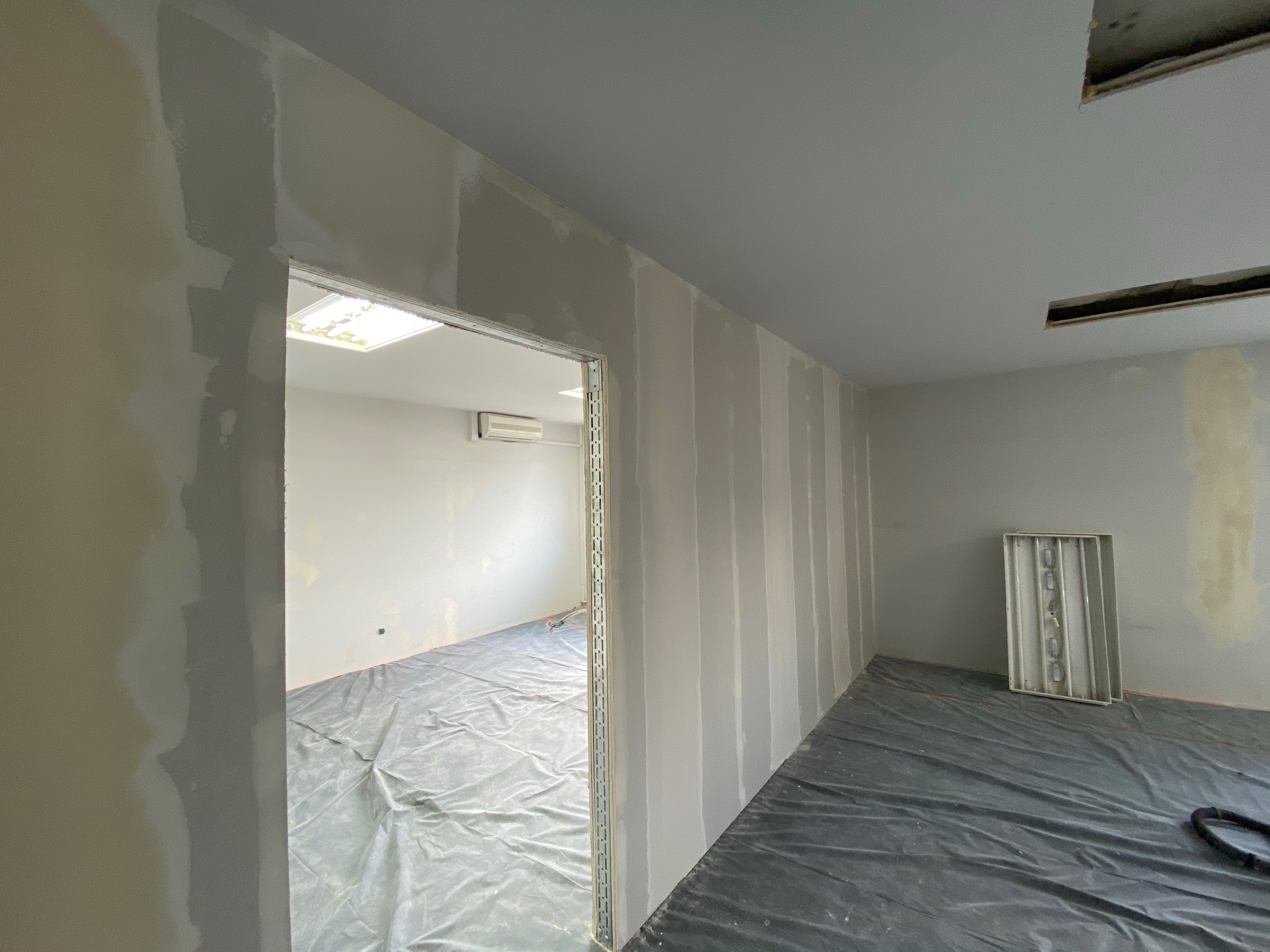 Wnętrze w trakcie prac wykończeniowych: pionowe pasy świeżo nałożonej masy na ścianach z płyt kartonowo-gipsowych, podłoga zabezpieczona folią malarską, widoczna rama drzwiowa i fragment sufitu...