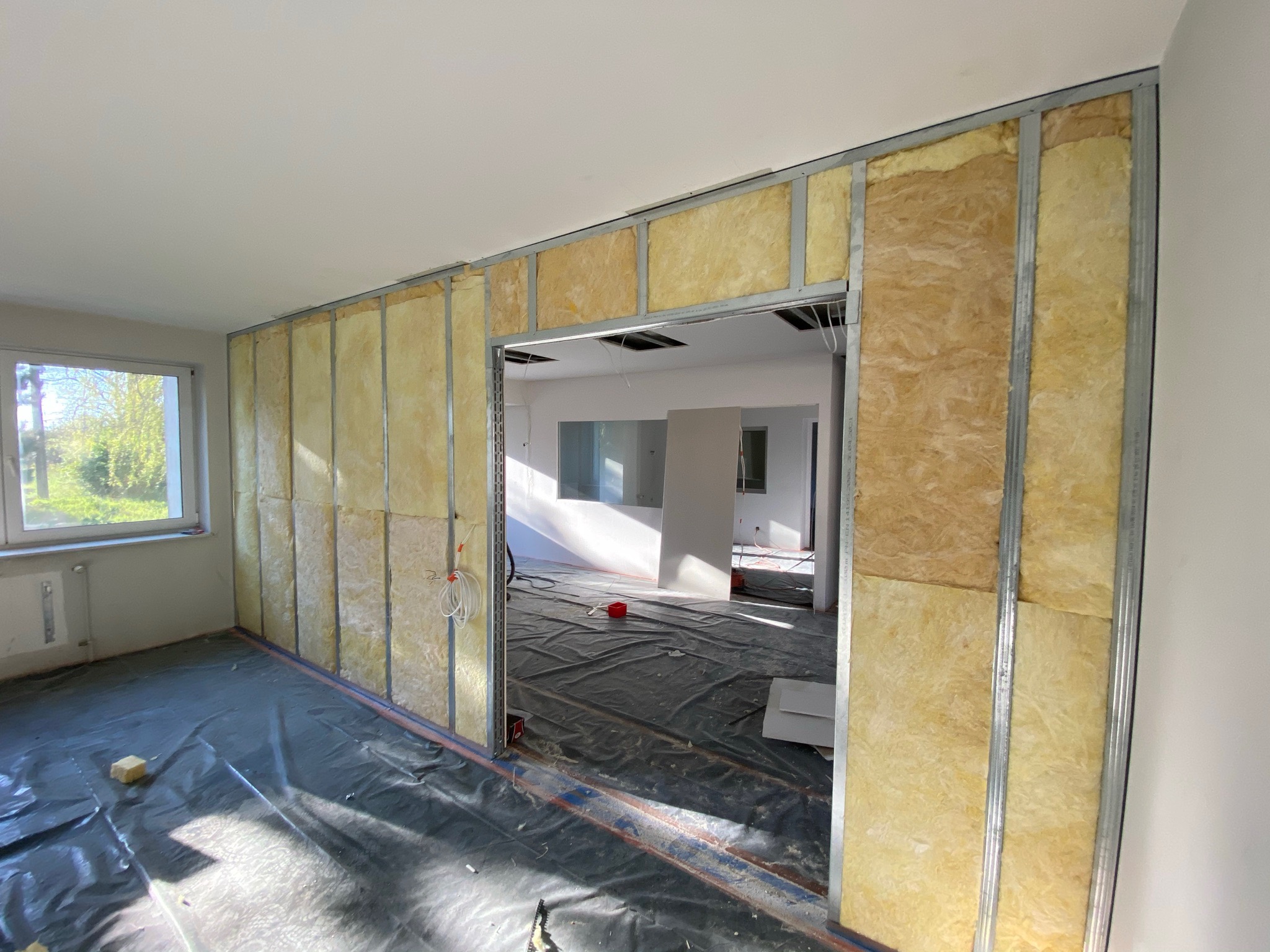 Wnętrze w trakcie prac: ściana z metalowym stelażem wypełnionym wełną mineralną, widoczne przewody elektryczne, podłoga zabezpieczona folią, fragment okna w tle.