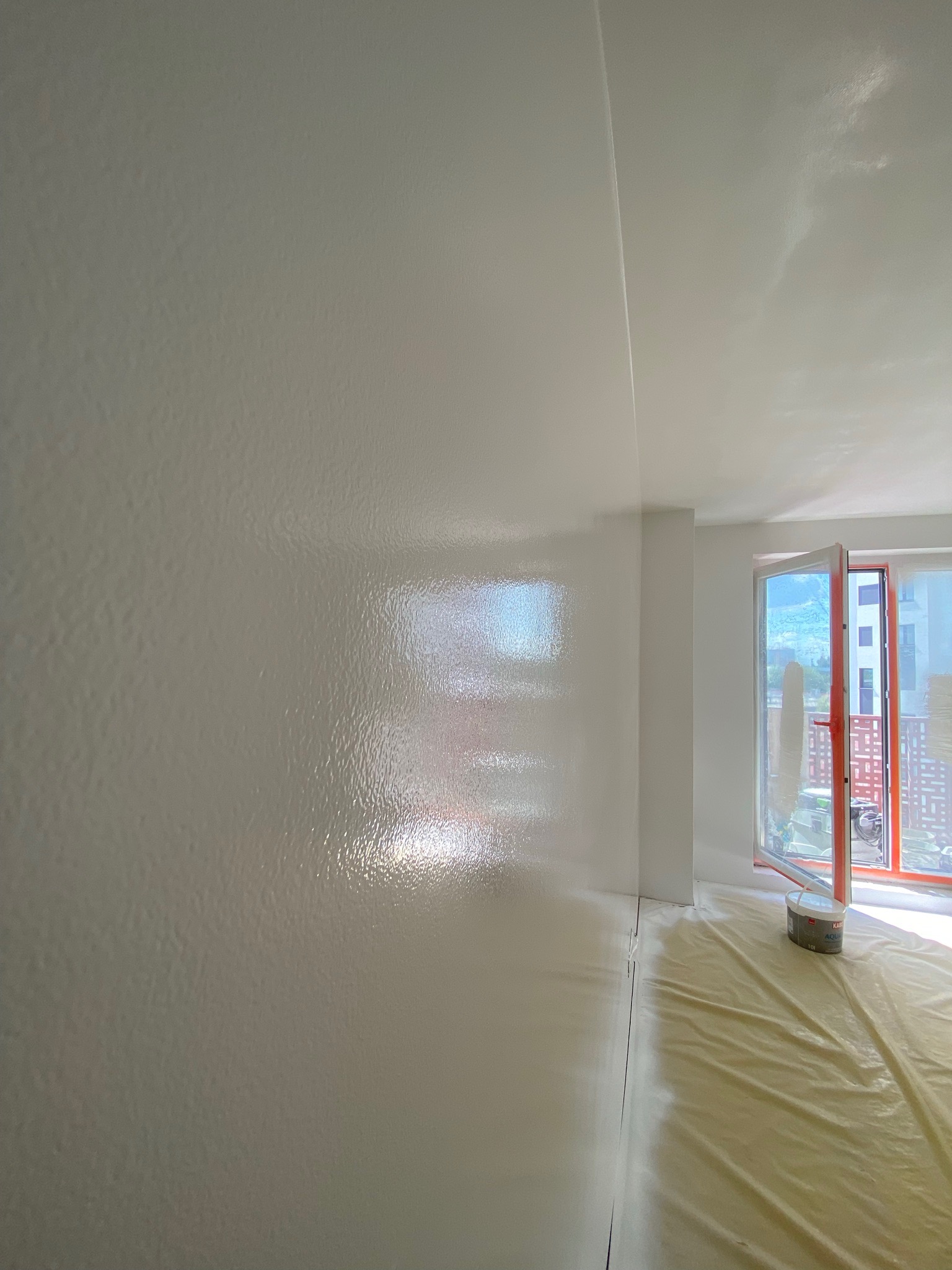 Świeżo pomalowane na biało wnętrze z widoczną fakturą na ścianie, zabezpieczona folią podłoga, otwarte okno balkonowe wpuszczające jasne światło.