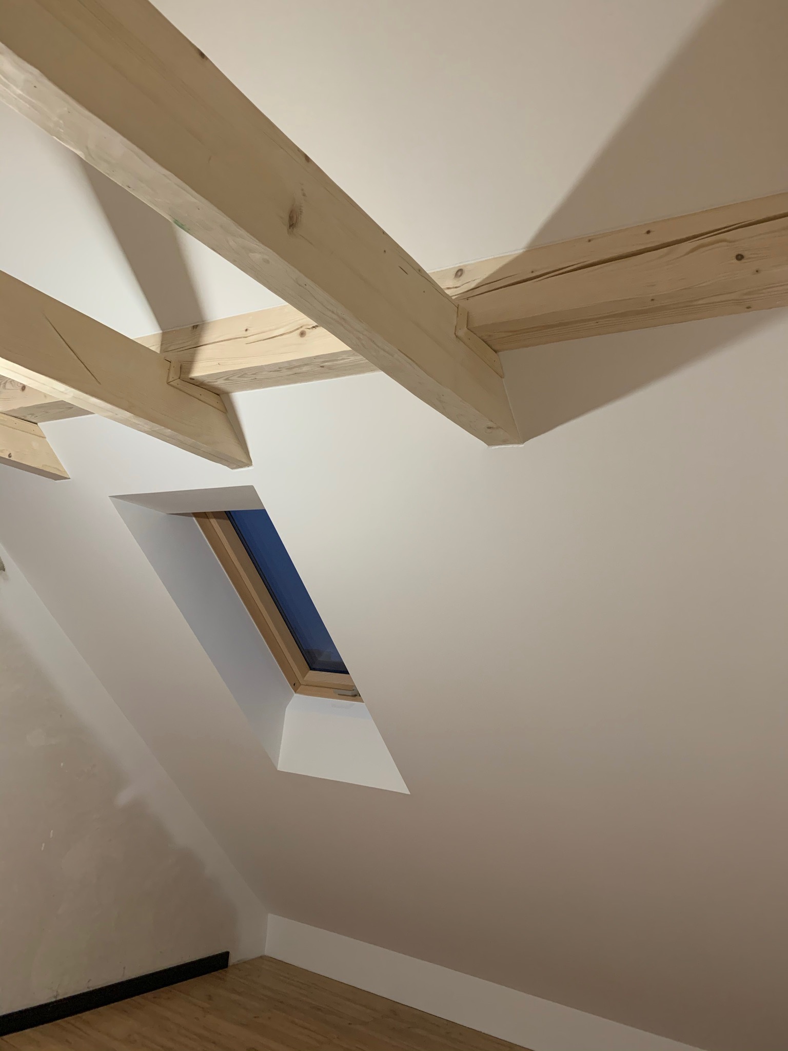 Poddasze po remoncie: drewniane belki stropowe, okno dachowe w wykończonej wnęce i świeżo pomalowane ściany w odcieniach bieli.