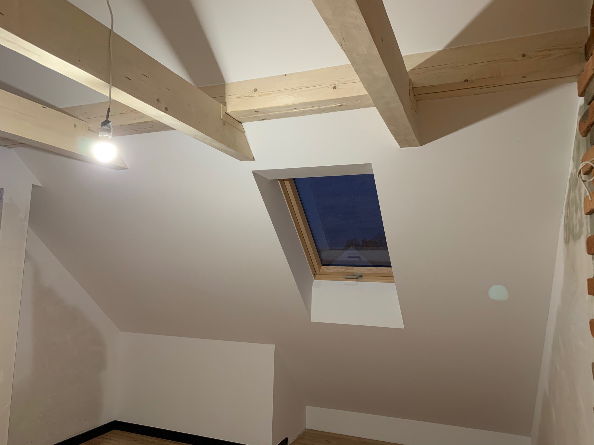 Poddasze z oknem dachowym po remoncie, świeżo pomalowane na biało, widoczne drewniane belki stropowe i fragment muru z cegły po prawej stronie. Jedna żarówka zawieszona na kablu oświetla przestrzeń.