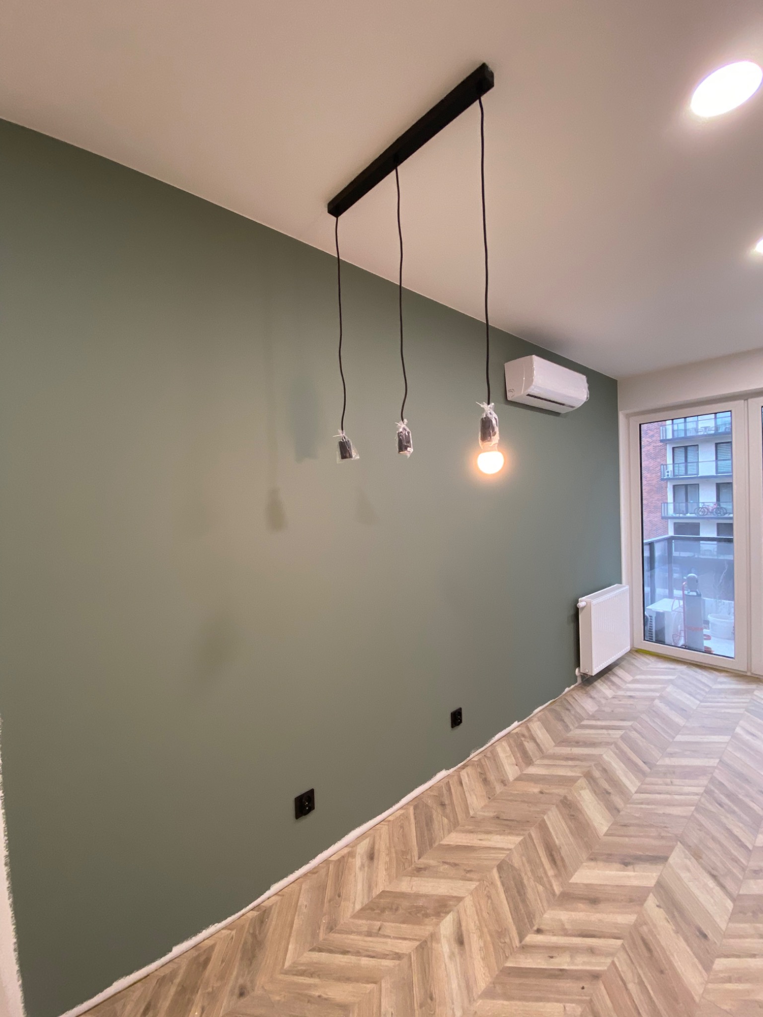 Pomieszczenie po malowaniu ścian na oliwkowy kolor, widoczne niedokończone oświetlenie z trzema żarówkami, grzejnik, klimatyzator i parkiet ułożony w jodełkę.