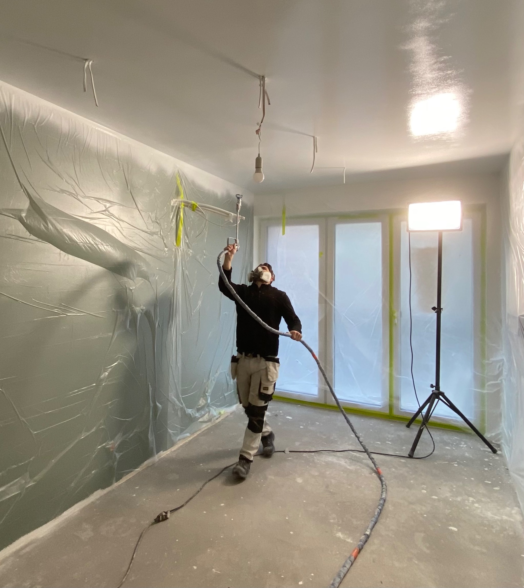 Pracownik w masce ochronnej aplikuje farbę natryskową w pomieszczeniu zabezpieczonym folią malarską, widoczna lampa na statywie oświetlająca ścianę.