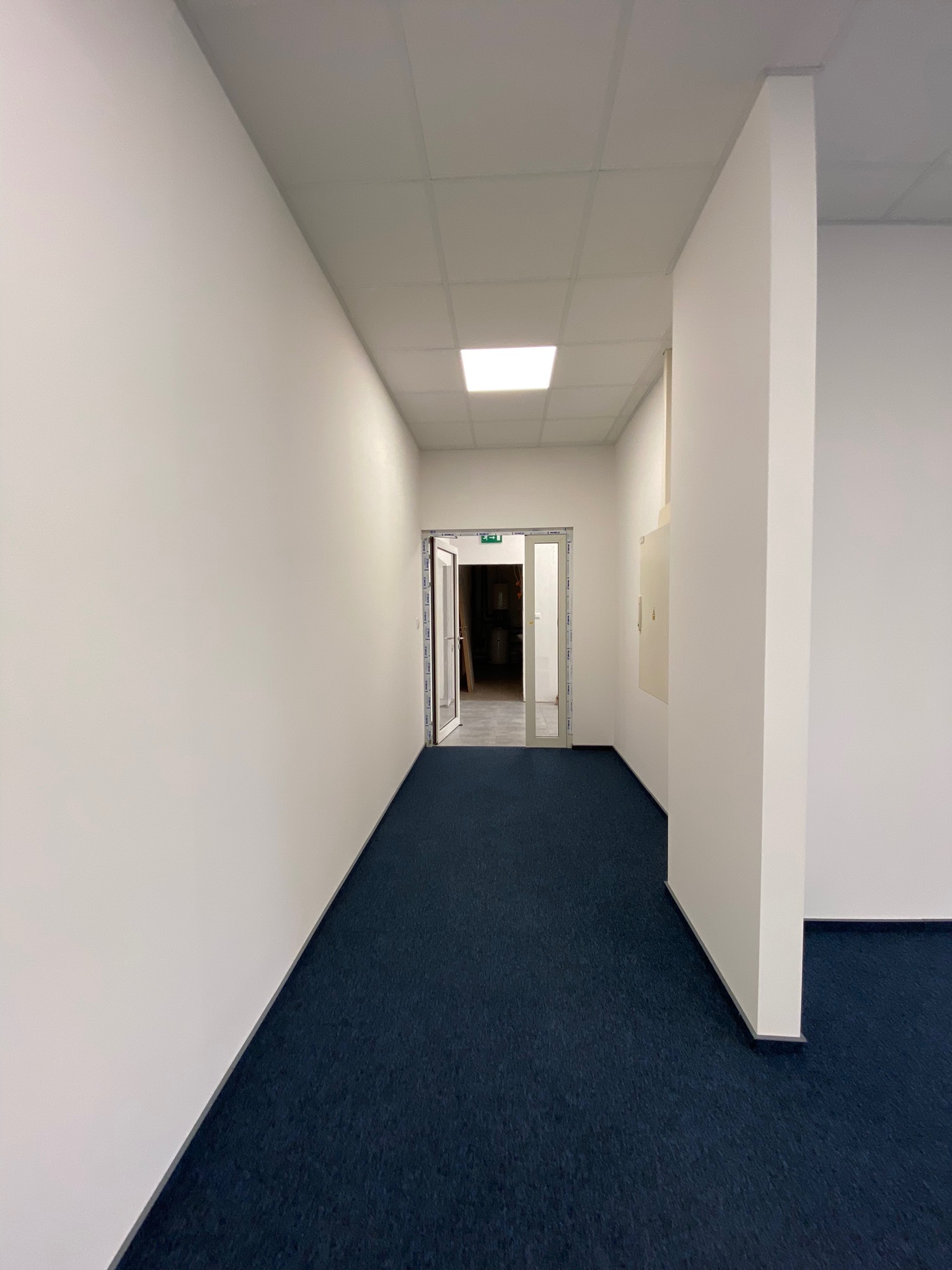 Korytarz biurowy z białymi ścianami, ciemnoniebieską wykładziną i otwartymi szklanymi drzwiami na końcu.
