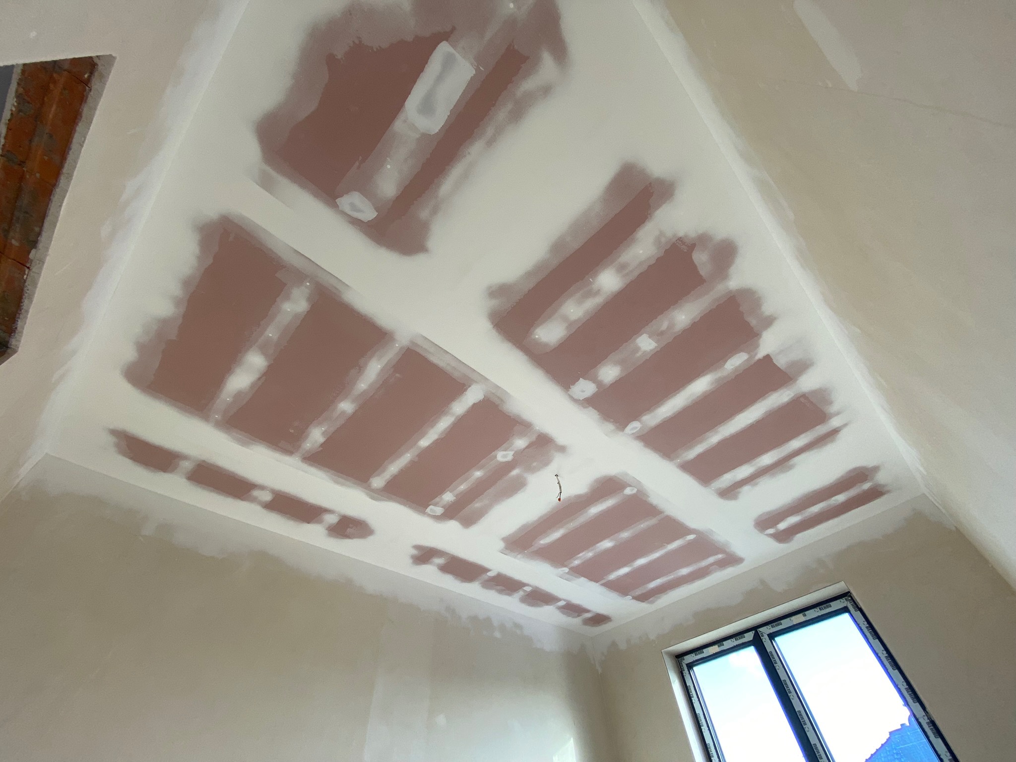 Sufit z widocznymi płytami kartonowo-gipsowymi, częściowo pokrytymi białą masą szpachlową, w trakcie prac wykończeniowych. W tle widoczne okno z niebieskim niebem.