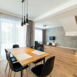 Jasne, przestronne wnętrze domu z drewnianą podłogą, stołem jadalnym z czarnymi krzesłami, schodami z podświetleniem LED i dekoracyjną ścianą z efektem betonu.