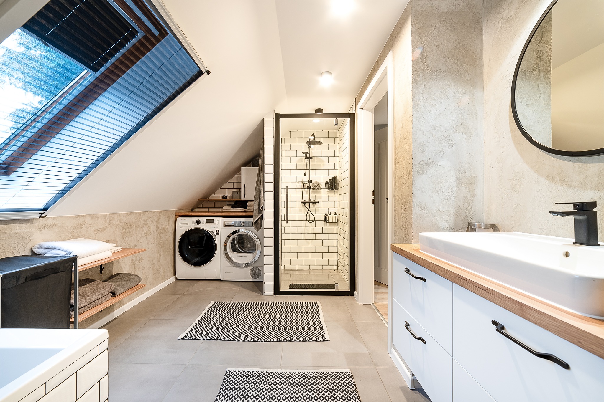Jasna łazienka na poddaszu z kabiną prysznicową w czarnej ramie, pralką i suszarką obok, okrągłym lustrem i białą umywalką z czarną armaturą.