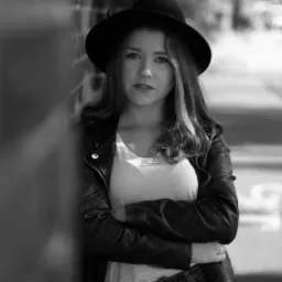 Portret młodej kobiety w czarnym kapeluszu i skórzanej kurtce, oparta o ceglaną ścianę, czarno-białe zdjęcie z rozmytym tłem miejskim.