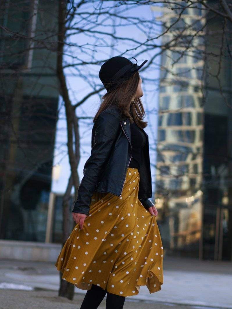 Kobieta w czarnej kurtce i musztardowej spódnicy w białe kropki pozuje na tle nowoczesnej architektury miejskiej, ubrana w kapelusz.