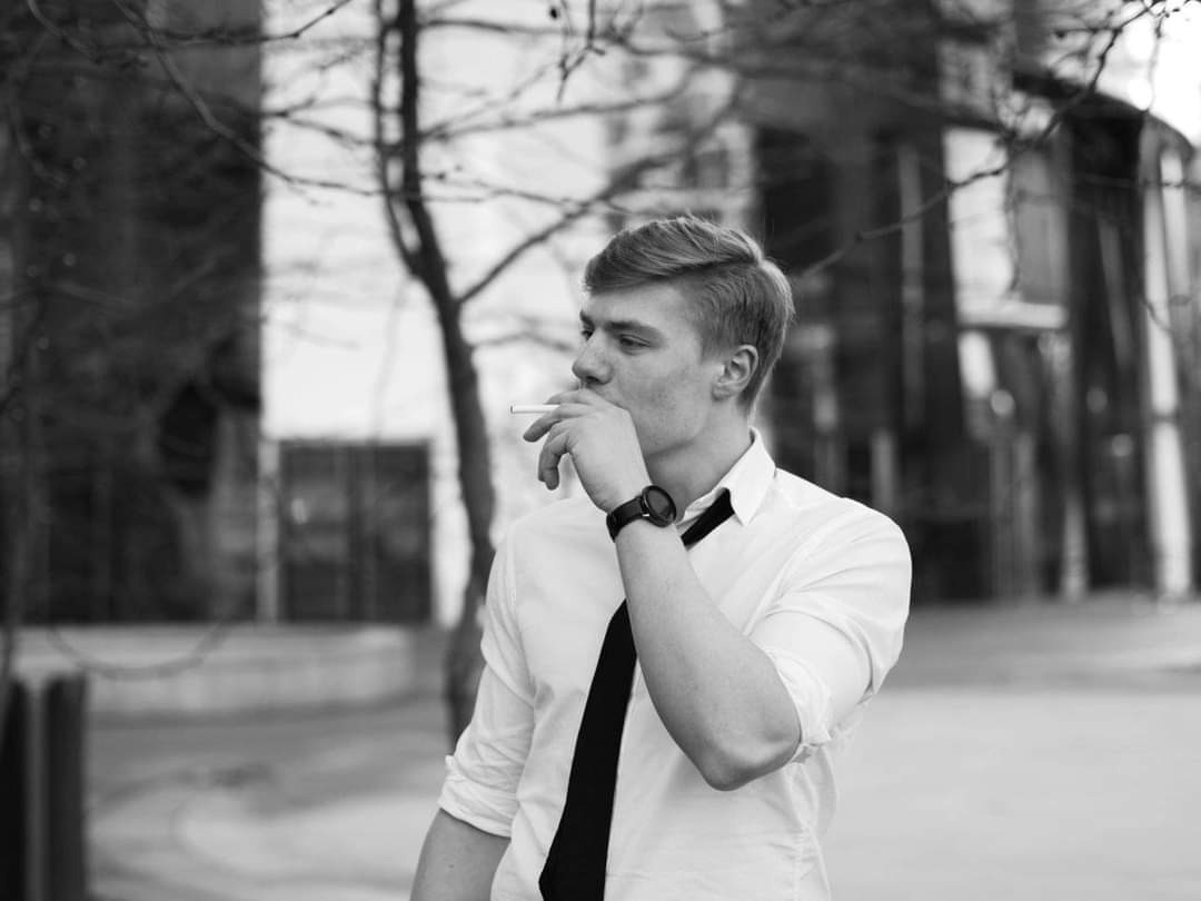 Czarno-białe zdjęcie młodego mężczyzny w koszuli i krawacie, palącego papierosa na tle miejskiego krajobrazu z drzewami i budynkami.