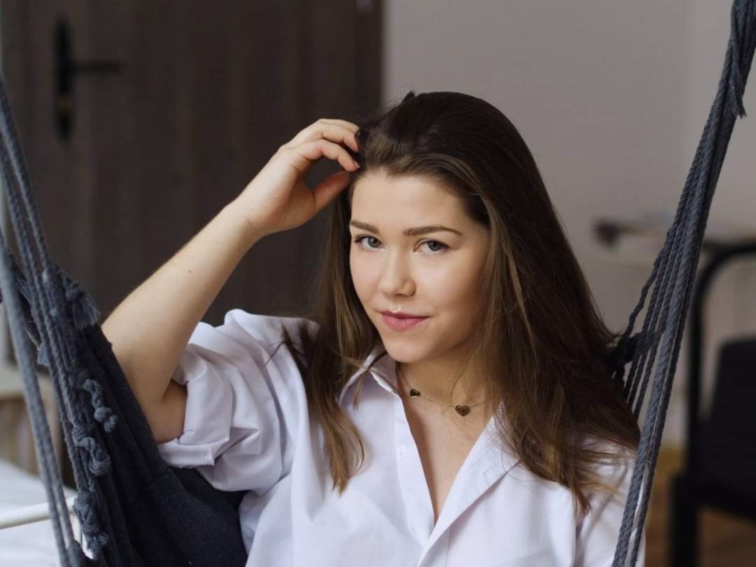 Portret młodej kobiety w białej koszuli, siedzącej w szarym hamaku linowym, z delikatnym naszyjnikiem w kształcie serca.