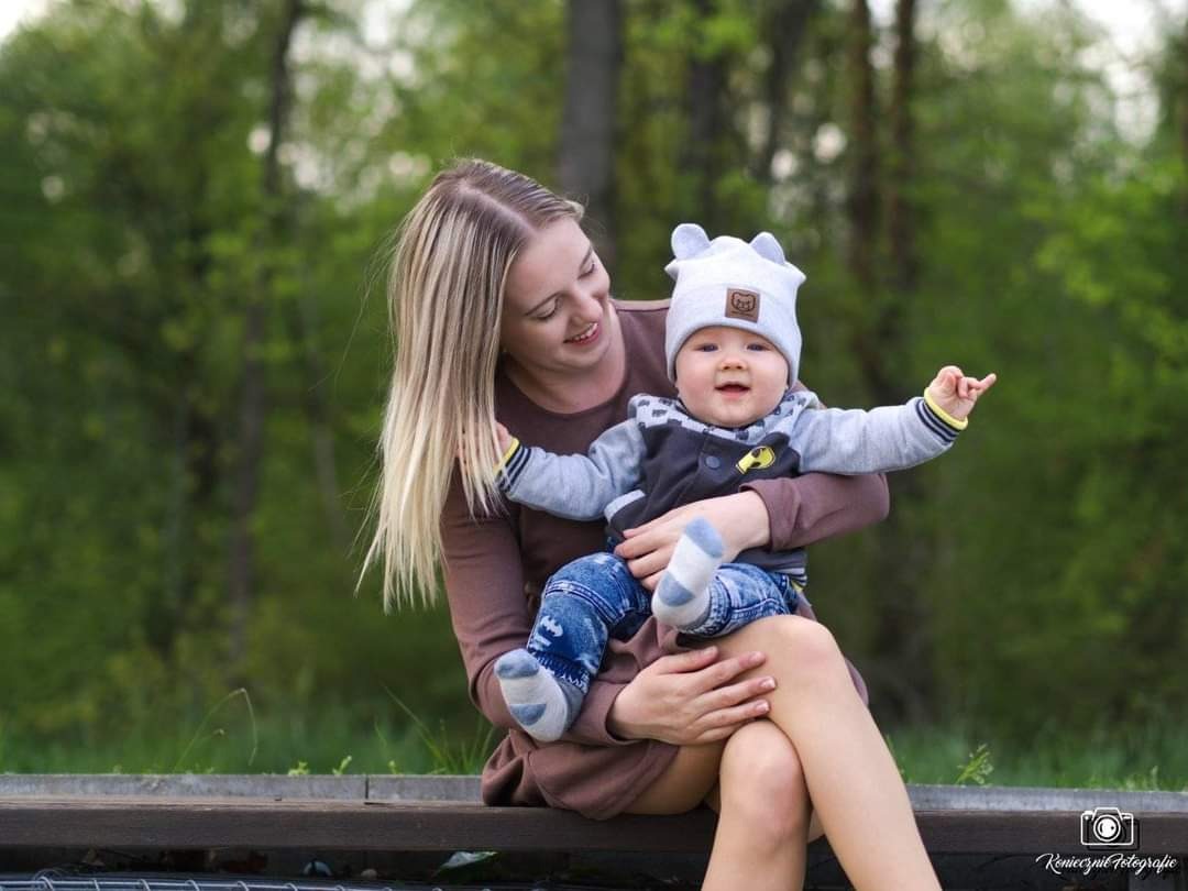 Uśmiechnięta blondynka trzyma na kolanach roześmianego niemowlaka w czapce z uszami na tle zieleni parku.