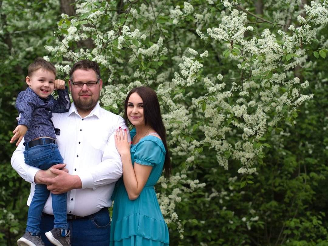 Uśmiechnięta rodzina: tata trzyma synka, mama stoi obok na tle kwitnących drzew, jasne, naturalne światło.