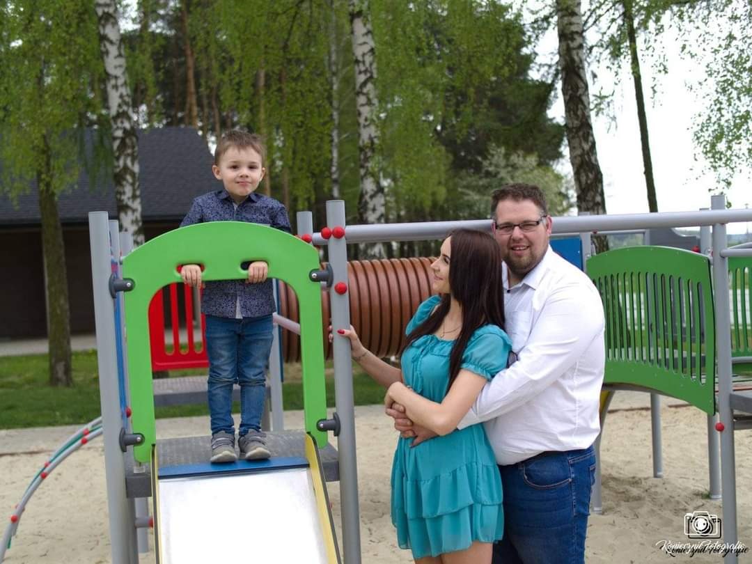 Rodzina pozuje na placu zabaw: chłopiec stoi na zjeżdżalni, rodzice obok, jasne ubrania, drzewa w tle.