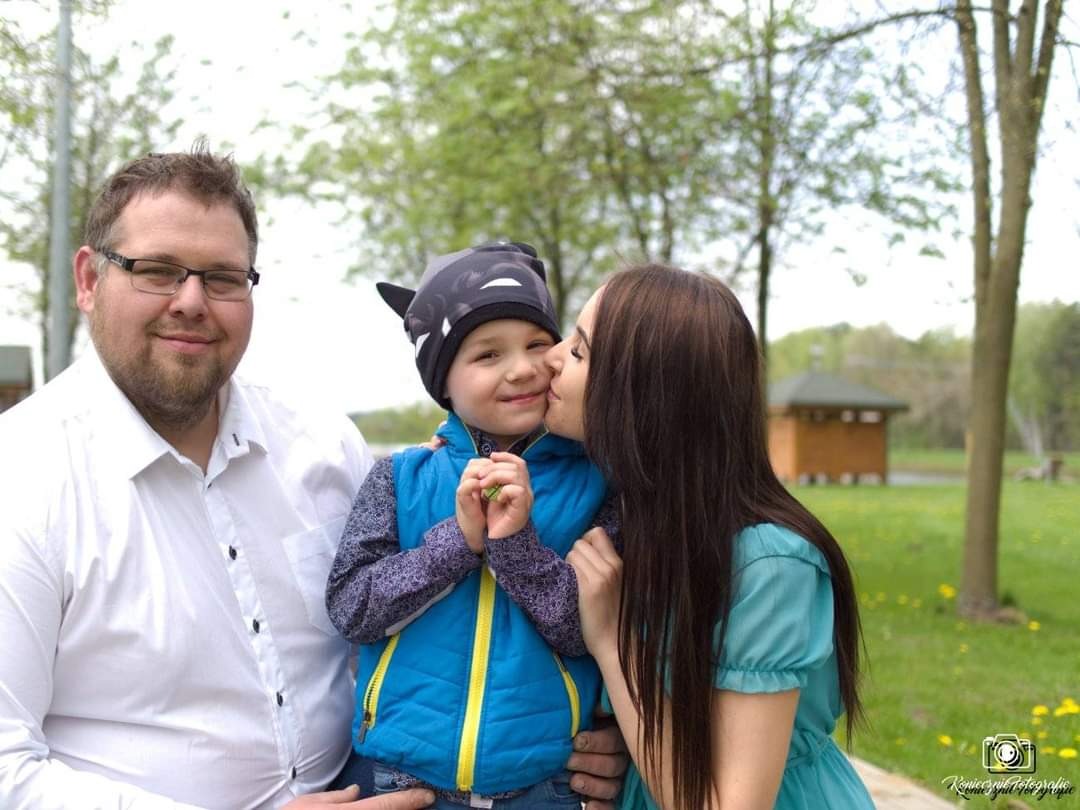 Uśmiechnięta rodzina na tle parku: ojciec w białej koszuli, syn w czapce z uszami i niebieskiej kamizelce, matka całująca syna w policzek, wiosenna sceneria.