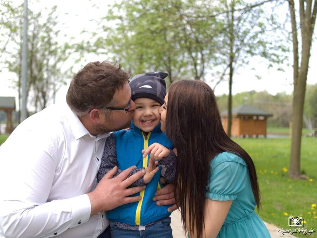 Uśmiechnięty chłopiec w niebieskiej kurtce i czapce z daszkiem całowany w policzki przez rodziców na tle zielonego parku.